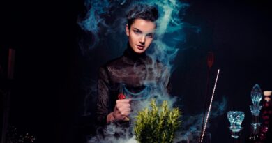 Graceful young female alchemist with knife in hand in black outfit preparing potion from various herbs among smoke in dark room